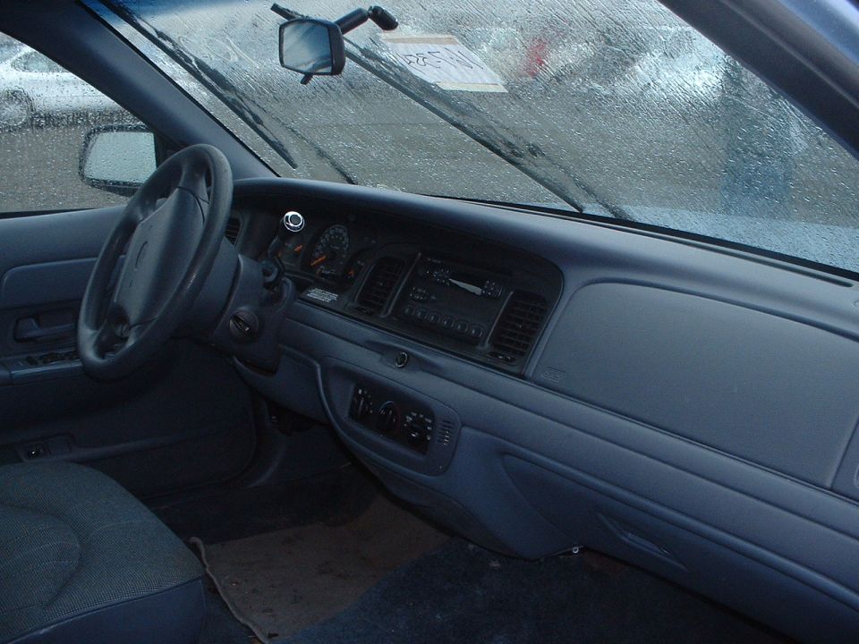 Interior of a 1999 Ford Crown Victoria, showing the dashboard, steering wheel, and seats. The windshield has water droplets