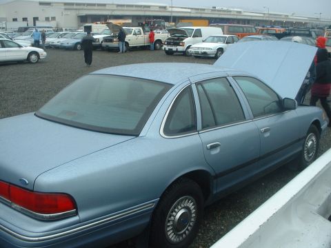 1999 Ford Crown Victoria - A light blue 1999 Ford Crown Victoria is shown from the rear quarter angle in a lot with other vehicles.