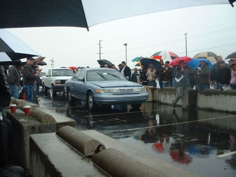 1999 Ford Crown Victoria - A blue sedan is displayed at an outdoor auction on a rainy day, with spectators holding umbrellas.