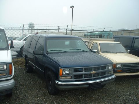 1999 Ford Crown Victoria - Several vehicles are parked in an outdoor lot under a cloudy sky, with a fence and water tower in the background.