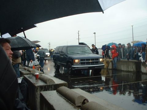 1999 Ford Crown Victoria - A dark blue SUV is driven through a wet auction lot as onlookers hold umbrellas.