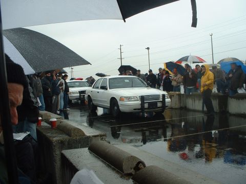 1999 Ford Crown Victoria - A white police car is displayed at an outdoor auction on a rainy day, with spectators holding umbrellas.