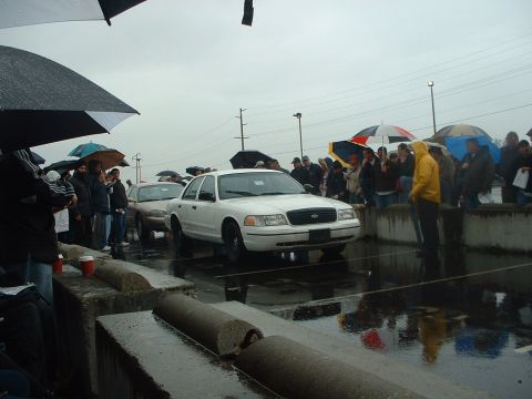 1999 Ford Crown Victoria - A white Ford Crown Victoria is displayed at an auction on a rainy day, with a crowd gathered under umbrellas.