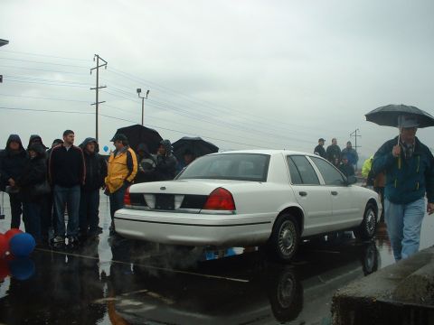 1999 Ford Crown Victoria - A white sedan is parked on a wet surface while people gather around holding umbrellas.