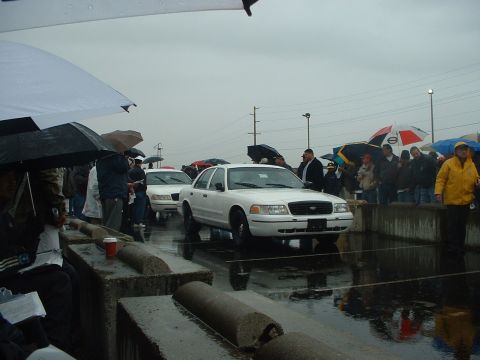 1999 Ford Crown Victoria - A white Ford Crown Victoria is presented at an outdoor auction on a rainy day, with several other vehicles and onlookers visible.