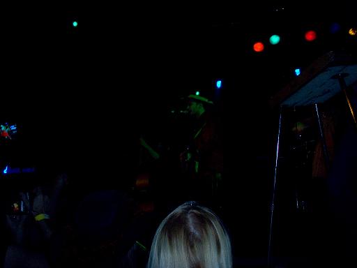 Musician playing guitar on a dark stage with colorful lights overhead. Audience in foreground
