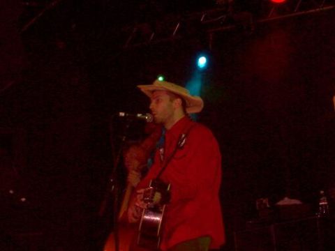 1988 Ford Bronco - A performer with a guitar and a cowboy hat sings into a microphone on a dimly lit stage with colored lights.