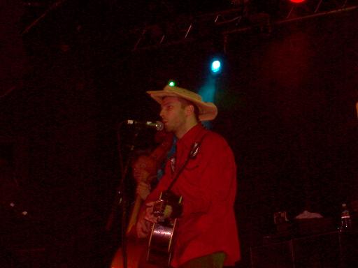 Man in cowboy hat and red shirt plays guitar and sings into a microphone on a dark stage
