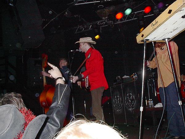 Musicians on a dimly lit stage, one in a red shirt and hat plays guitar, another plays a double bass, and a third plays a keyboard