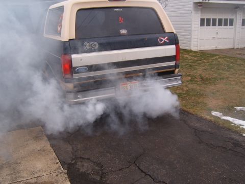 1993 Ford Bronco - Rear view of a two-tone tan-over-dark-green Ford Bronco full-size SUV billowing thick white exhaust smoke in a cracked asphalt driveway. Th…