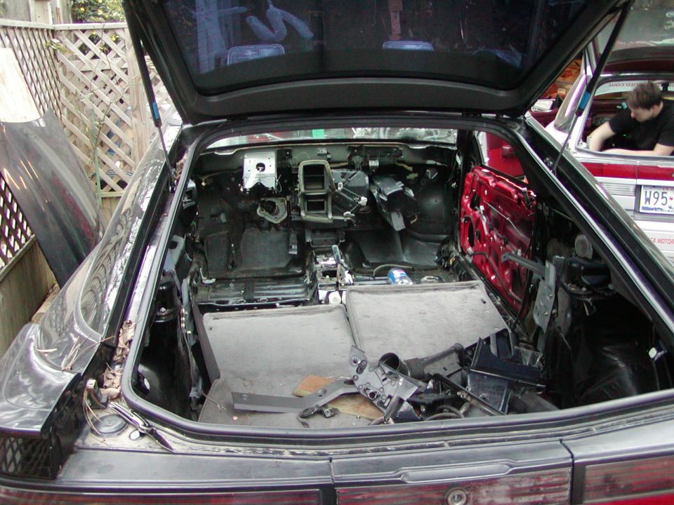 Black hatchback with rear hatch open, interior fully stripped to bare metal, red door and parts inside