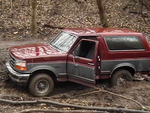 1986 Ford Bronco - A maroon and gray Ford Bronco is stuck in a muddy, wooded area with its driver's door ajar.