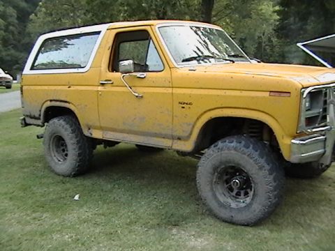 1986 Ford Bronco - A muddy yellow Ford Bronco with a white top sits on grassy terrain.