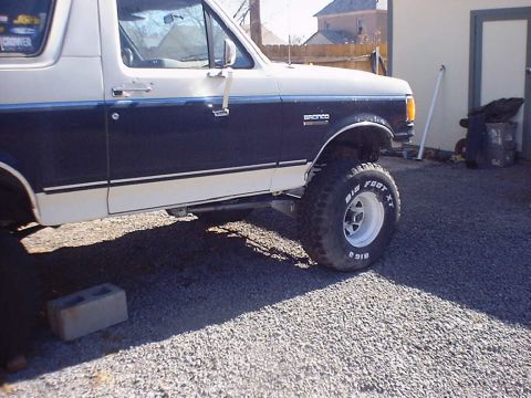 1988 Ford Bronco - A white and dark blue Ford Bronco is parked on a gravel lot, showing its lifted suspension and large tires.