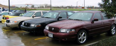 2000 Ford Crown Victoria - Several Ford Crown Victoria sedans and a yellow Ford Mustang convertible are parked in a lot on a rainy day.