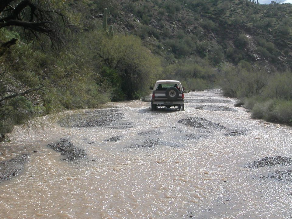 White Ford Bronco driving through a rocky, shallow stream in a desert landscape