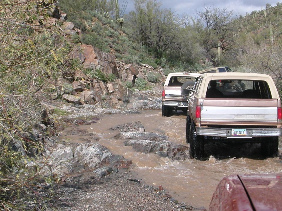 Rear view of a 1984 Ford Bronco driving through water on a rocky trail