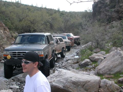 1984 Ford Bronco - Several modified SUVs navigate a challenging off-road path with large rocks and sparse vegetation.