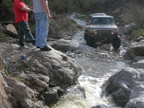 1984 Ford Bronco - Several trucks drive through a rocky riverbed, with two onlookers observing from the bank.