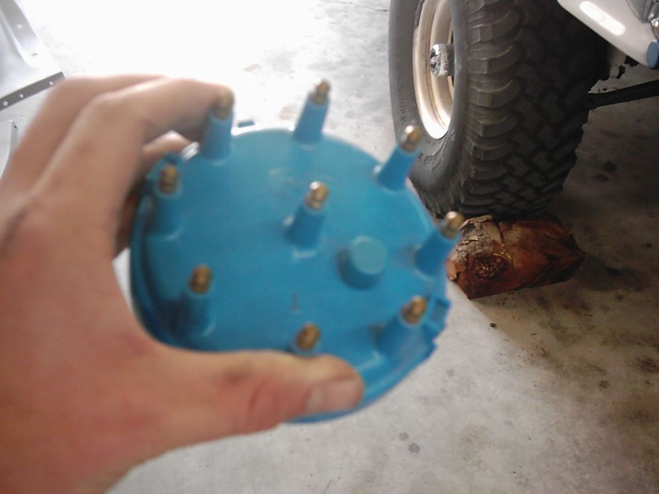 Hand holding a blue distributor cap with brass terminals. A tire and part of a vehicle are visible in the background