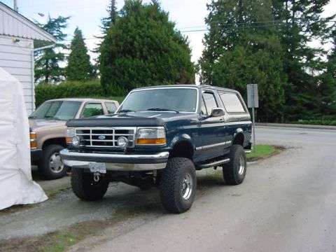 1993 Ford Bronco - A dark blue 1993 Ford Bronco is parked next to a tan SUV, a lifted suspension and prominent tires.