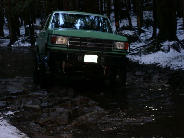 Green Ford pickup with brush guard crossing a rocky creek in snowy woods at night