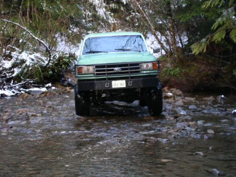 1978 Ford Bronco - Head-on front view of a lifted mint-green Ford truck fording a rocky creek, water up to the axles and mud-terrain tires. It wears a black t…