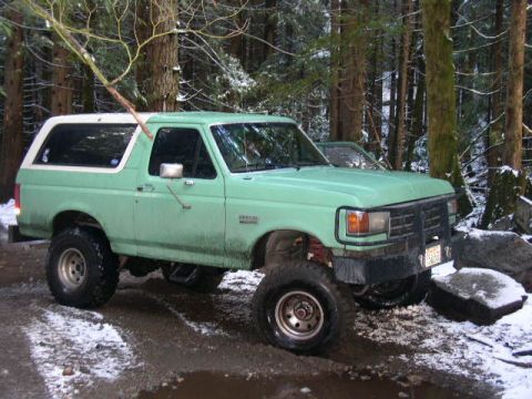 1978 Ford Bronco - Passenger-side three-quarter front view of a lifted mint-green full-size Ford Bronco with a white removable hardtop, parked on a snowy, mud…