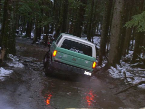 1978 Ford Bronco - A green early Ford Bronco with a white removable hardtop, seen from the rear as it climbs a muddy, water-filled forest trail with patches o…