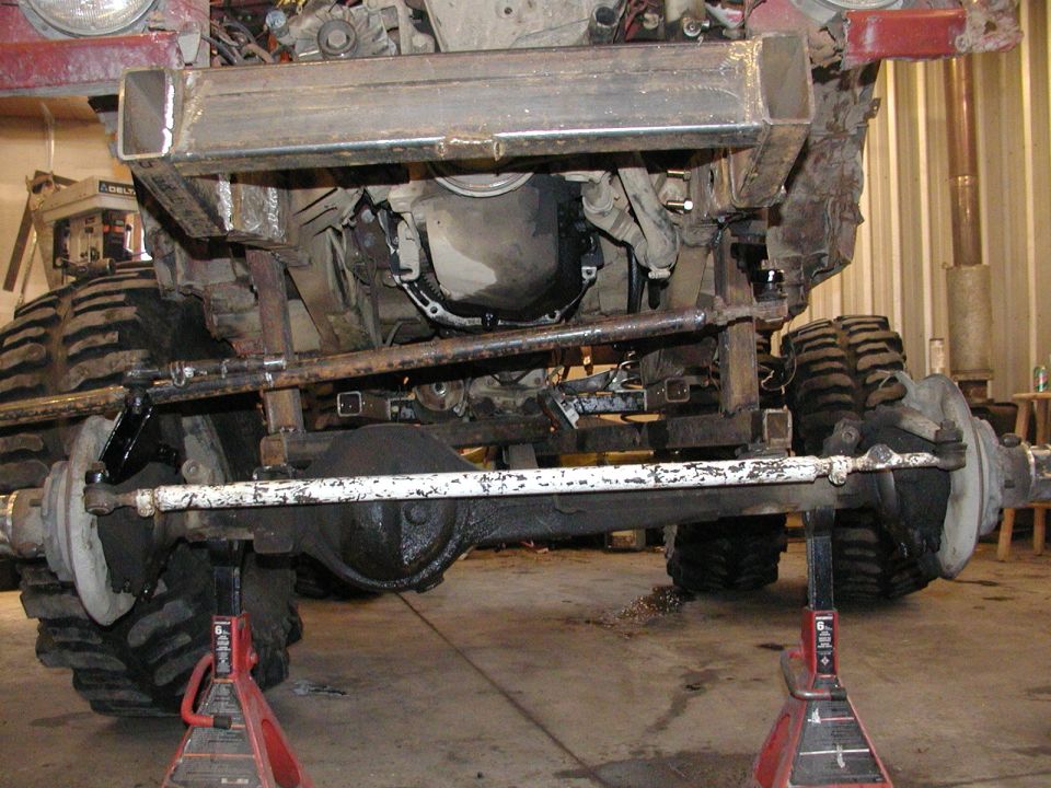 Solid front axle and white tie rod on a lifted 4x4 raised on red jack stands in a shop