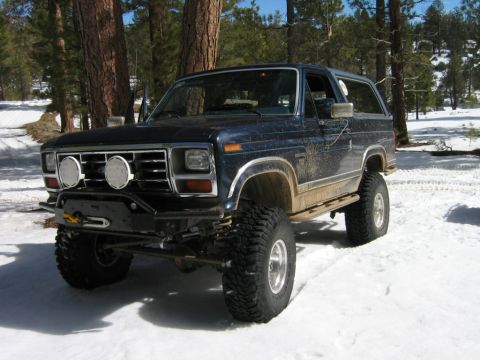 1985 Ford Bronco - A lifted Ford Bronco sits in a snowy, wooded area with its front end facing the camera.