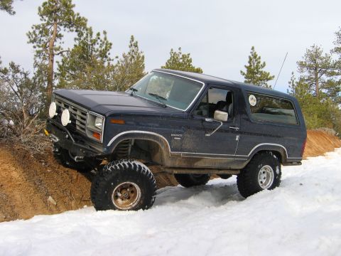 1985 Ford Bronco - A dark blue 1985 Ford Bronco navigates a snowy, off-road trail, its lifted suspension and tires.