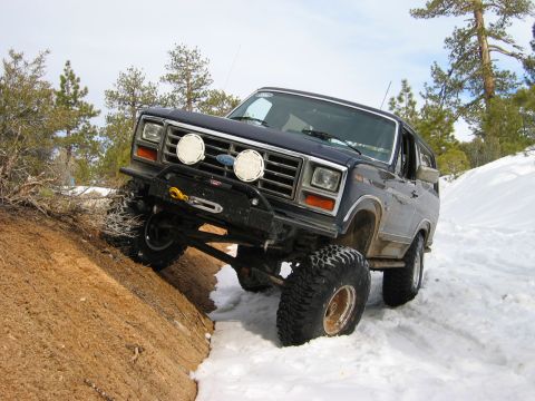 1985 Ford Bronco - Ford Bronco navigates a snowy, off-road trail with a steep dirt embankment.