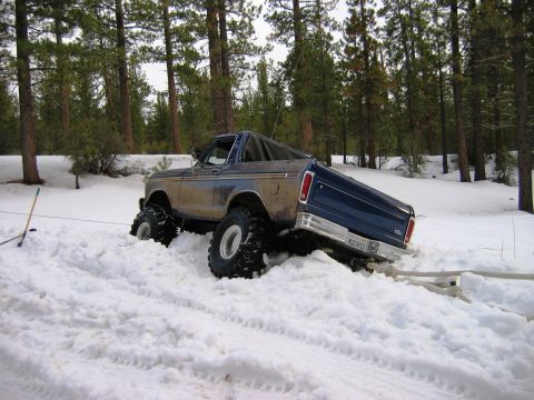 1985 Ford Bronco - A lifted Ford Bronco with large tires is stuck in deep snow, with trees in the background.