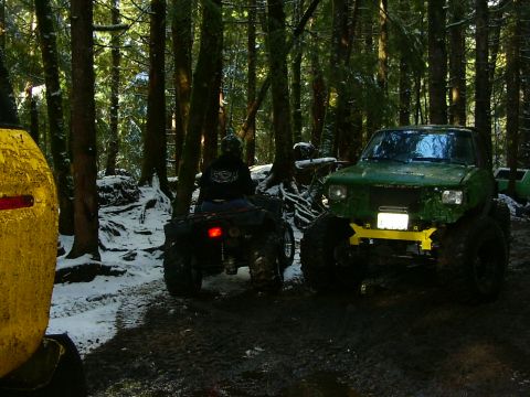 1990 Ford Bronco - A modified green off-road vehicle with a yellow bumper is parked in a muddy, snowy forest next to an ATV.