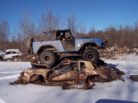 1974 Ford Bronco - A blue 1974 Ford Bronco is stacked on top of a severely damaged gold car in a snowy junkyard setting.