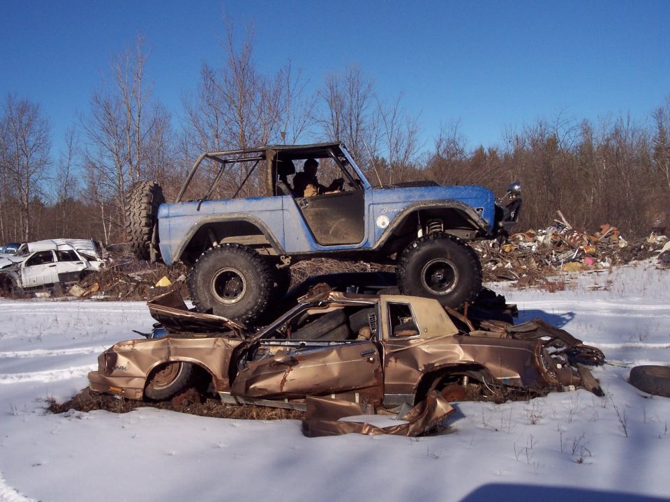 Blue 1974 Ford Bronco with roll cage, spare tire on rear, on top of a crushed gold car in a junkyard