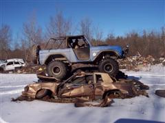 1974 Ford Bronco - A light blue Ford Bronco is perched atop two heavily damaged cars in a snowy, outdoor setting.