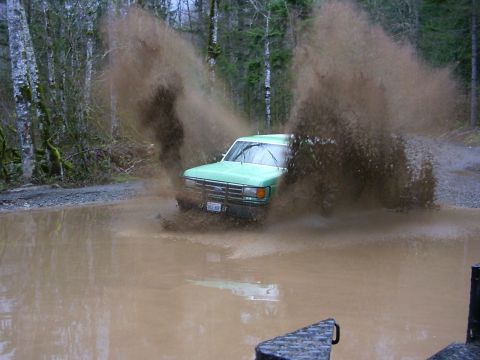 1990 Ford Bronco - A green 1990 Ford Bronco powers through a muddy puddle, sending water and dirt flying.
