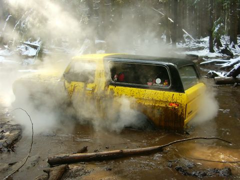 1990 Ford Bronco - A yellow SUV is stuck in a muddy, wooded area, with steam or smoke billowing around it.