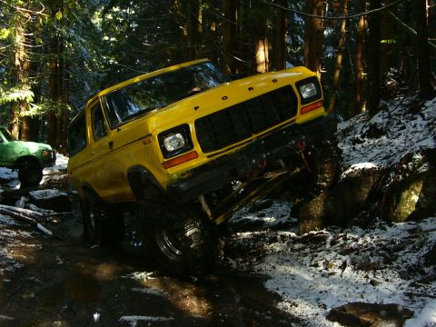 1990 Ford Bronco - Yellow Ford Bronco tackling an off-road trail in a snowy forest, with another vehicle following.