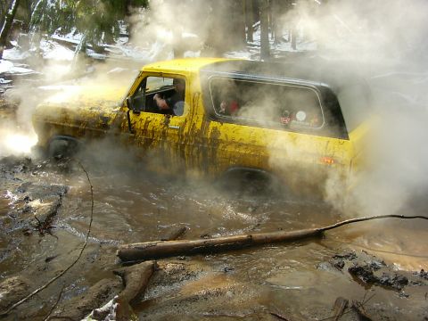 1990 Ford Bronco - This yellow Ford Bronco appears to be stuck deep in a muddy, wooded area, with steam or smoke billowing around it.
