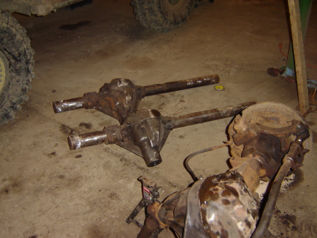 three rusty vehicle axles on a concrete floor, one in foreground has brake components attached