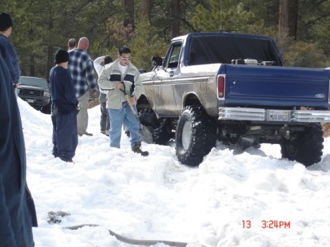 2000 Ford F-450 - A group of people are gathered around a lifted blue Ford pickup truck with large tires that appears to be stuck in the snow.