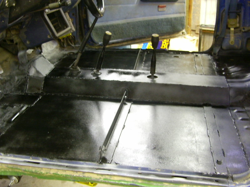 Vehicle interior floor with black protective coating applied, showing transmission tunnel and gear shifters