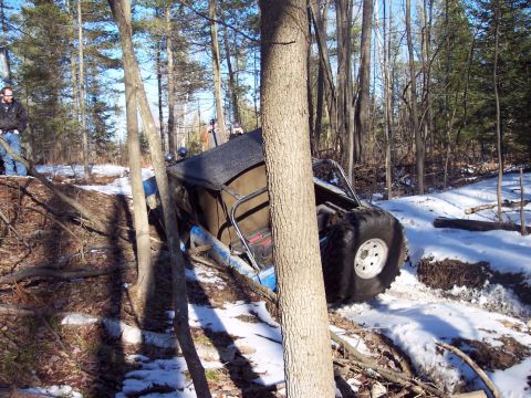 1974 Ford Bronco - An off-road vehicle is tilted precariously in a snowy wooded area, with large tires and a roll cage visible.