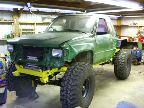 1990 Ford Bronco - Modified pickup truck with large tires and yellow frame, seen in a garage with visible tools and shelving.