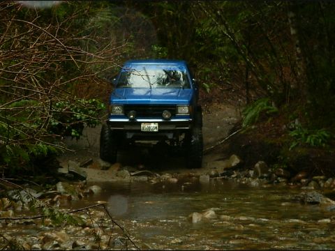 1990 Ford Bronco - A blue 1990 Ford Bronco navigates a rocky stream bed in a wooded area.