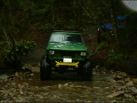 1990 Ford Bronco - A green off-road vehicle navigates a rocky stream bed surrounded by dense foliage.