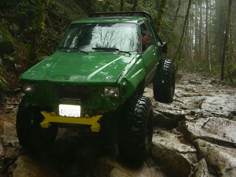 1990 Ford Bronco - This heavily modified off-road vehicle tackles a challenging rocky path through a dense, wet forest.